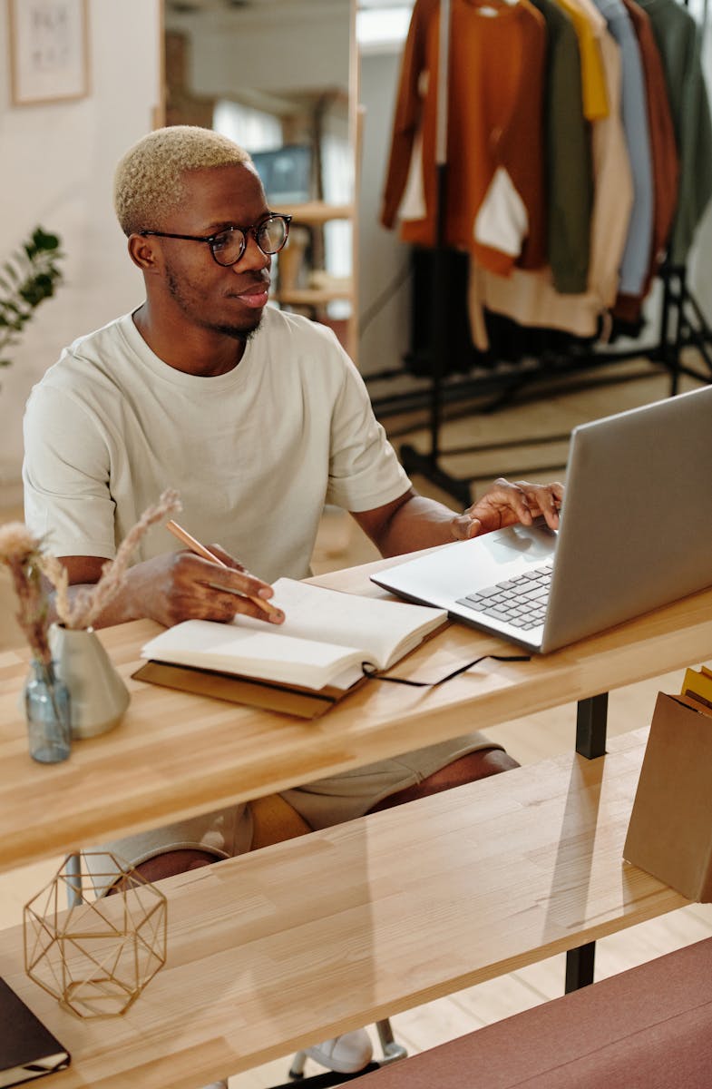 A man sitting at a desk writing in a notebook while using a laptop indoors, focused on work.