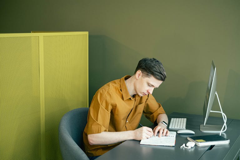 A man in a brown shirt sketching in a notebook at a tidy desk in a modern office setting.