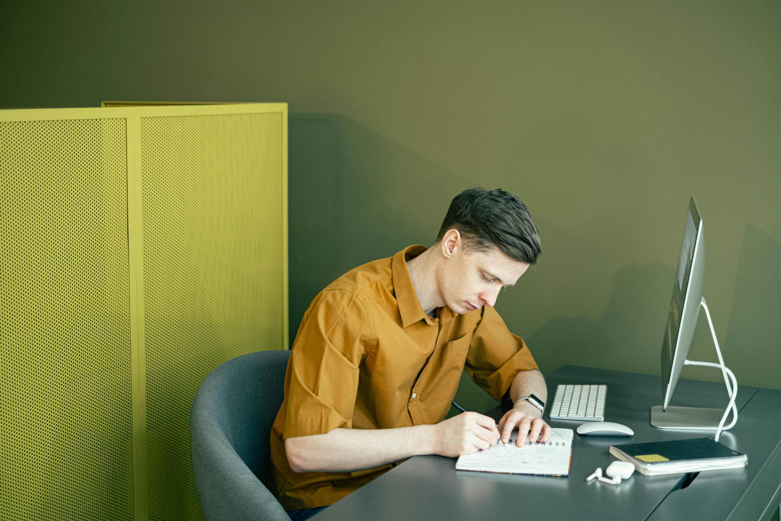 A man in a brown shirt sketching in a notebook at a tidy desk in a modern office setting.