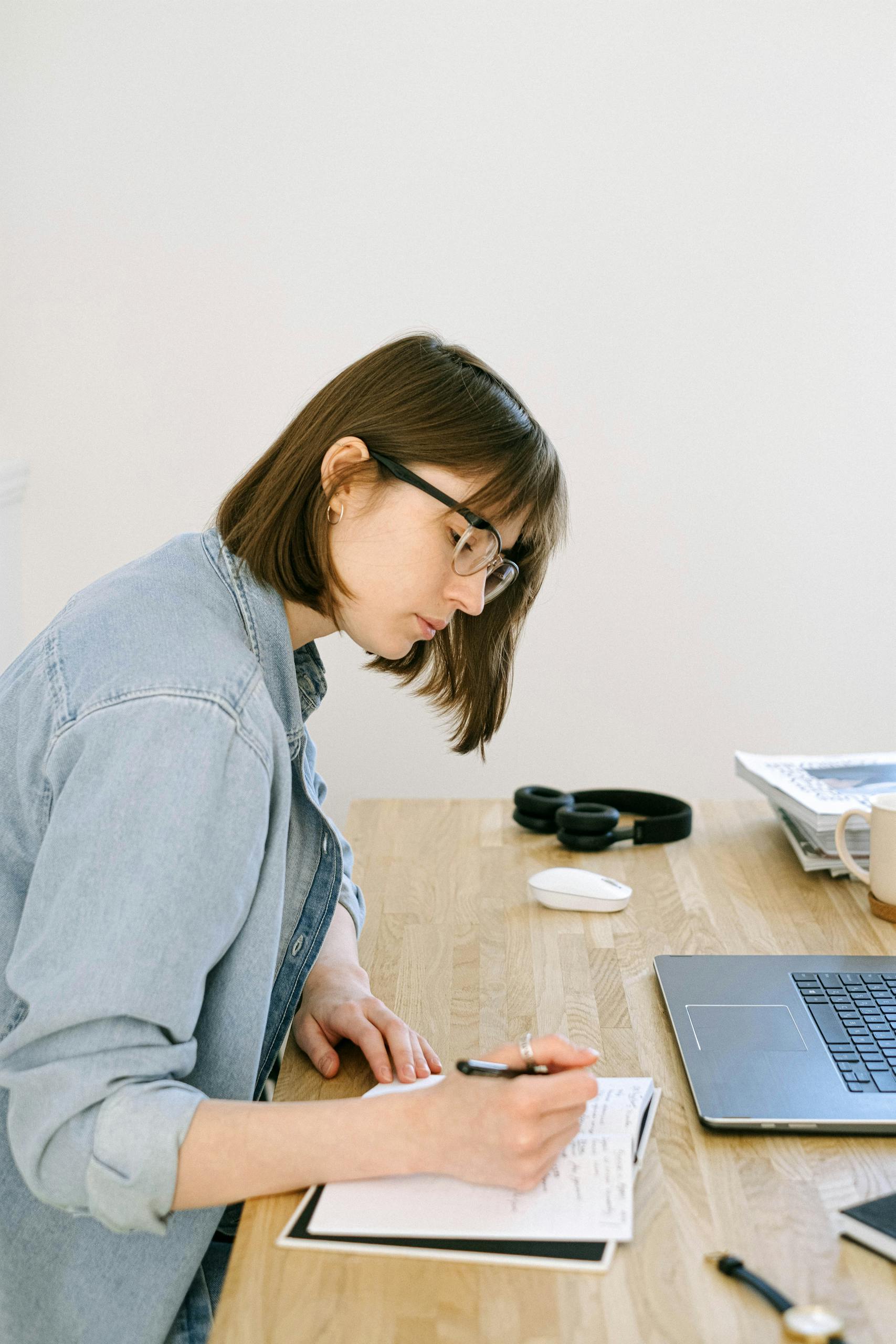 Focused young woman taking notes in a cozy home office setting with laptop and stationery.
