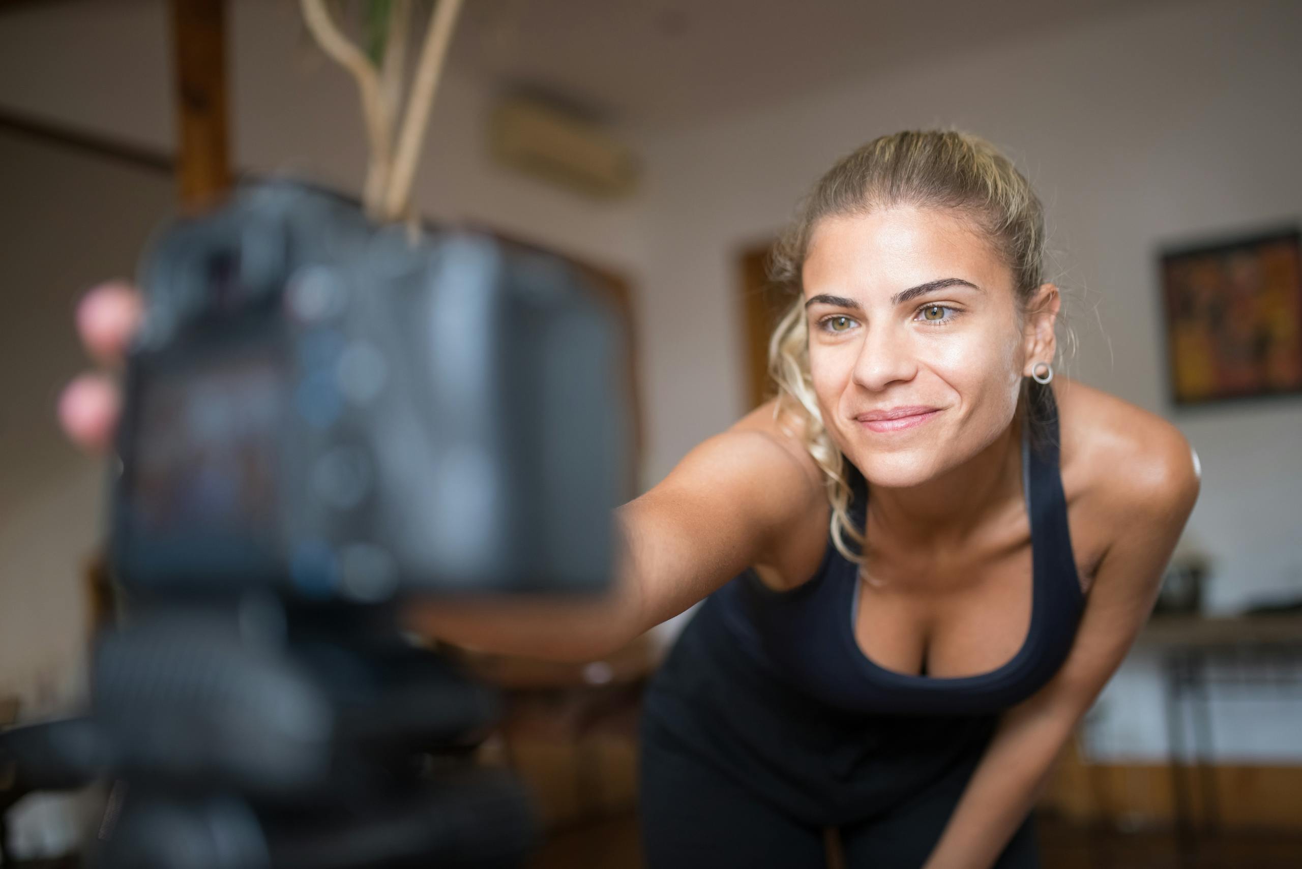 Smiling woman in sportswear setting up a camera indoors for a fitness recording session.