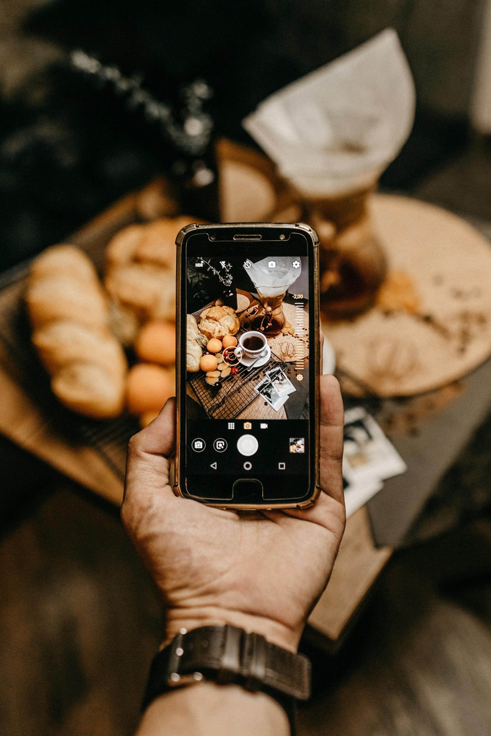 Warm indoor scene with smartphone capturing coffee and pastries on table.